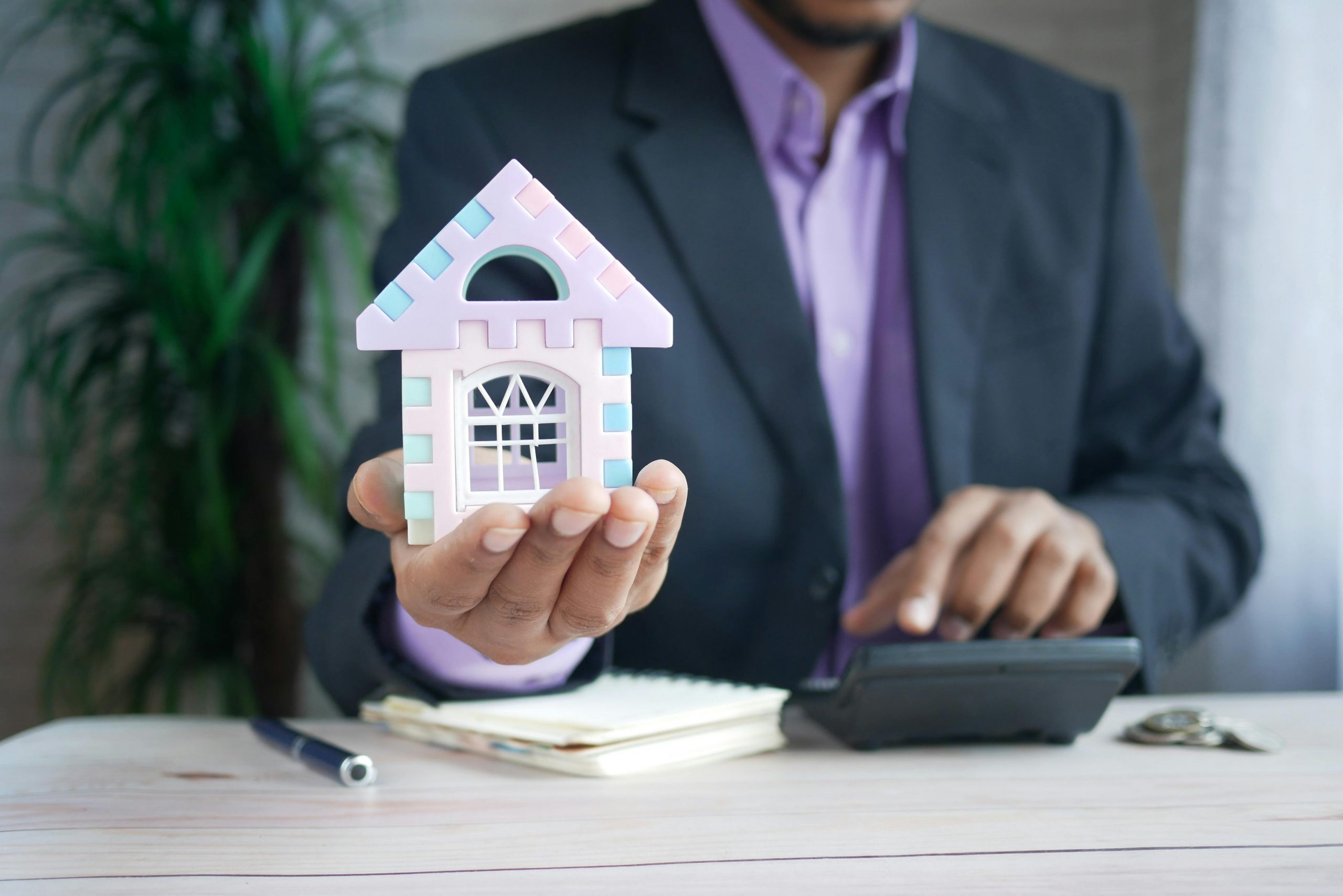 man in a suit holding a model house in one hand and using a calculator with the other